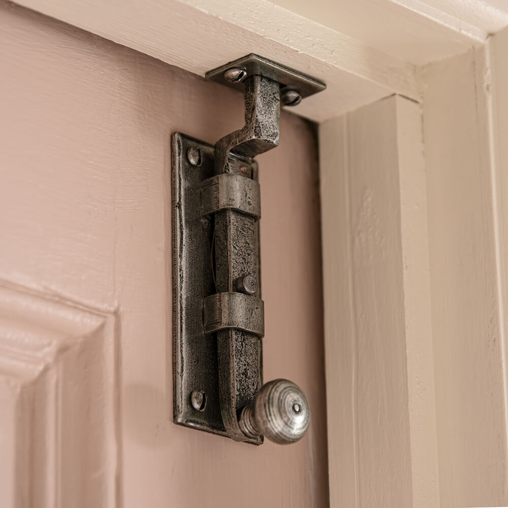 Pewter sliding knob bolt with a cranked design fitted to a light pink door that is secured to the top of the door frame