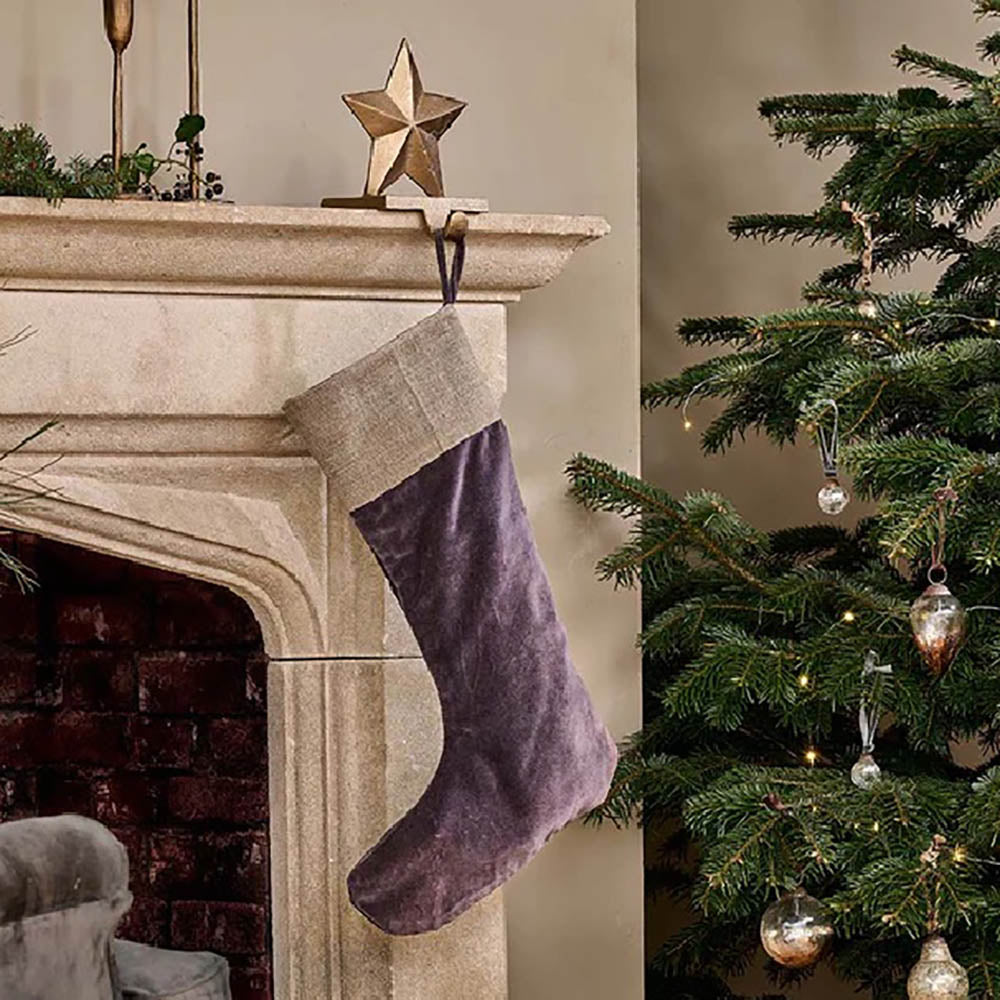 Charcoal velvet christmas stocking with jute band around the top. Hanging from stone fireplace on a gold star hook next to a christmas tree with gold baubles.