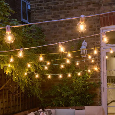 Pink, yellow and green festoon light hanging above a patio with a red brick wall and greenery in the background