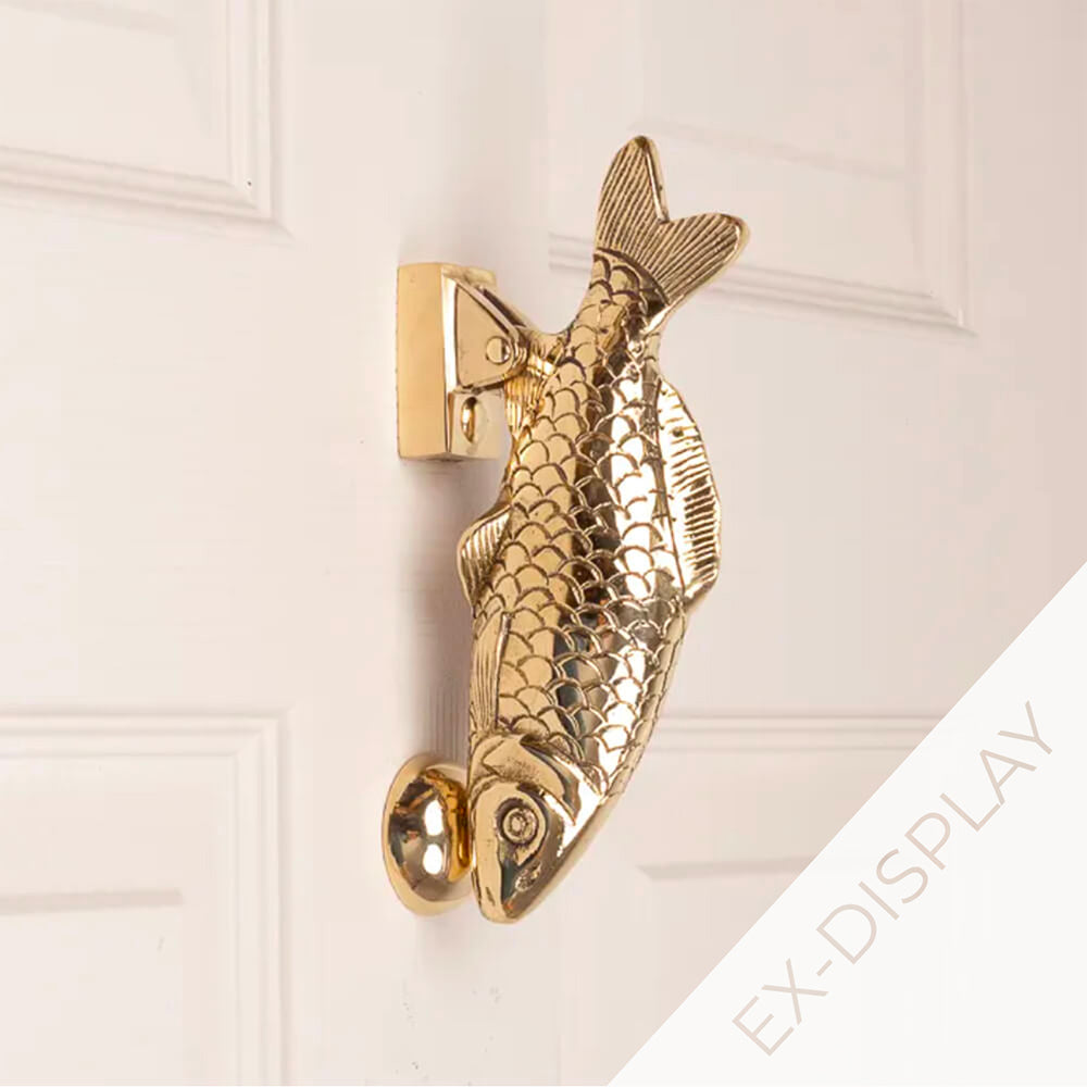 Solid brass carp door knocker with embossed scales and the face resting on the striker plate on a pale pink door with a watermark and ex display text in the corner