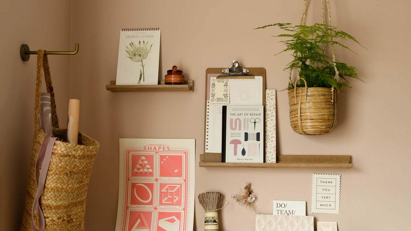 Corner of a room with dusky pinl walls, oak shelves and desk with lamp, plants, stationery on.