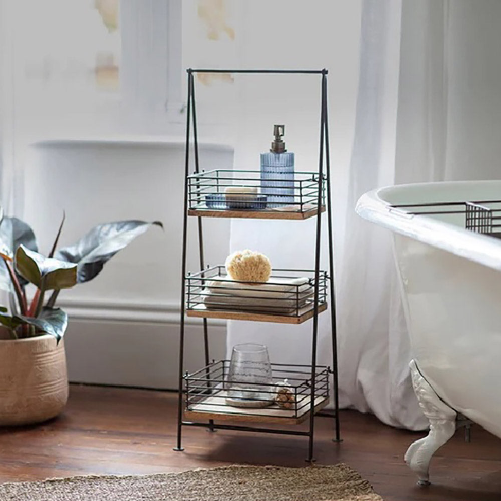 Three tier bathroom caddy with iron frame and mango wood shelves in bathroom setting with bathroom items on the shelves next to bath with window back drop