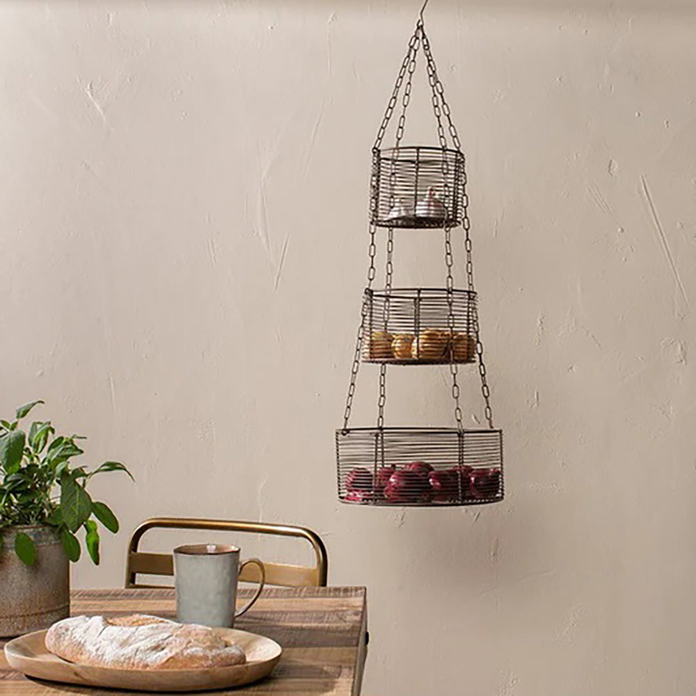 3 tier wire hanging baskets containing vegetables against taupe background next to a wooden table with loaf of bread and a green plant