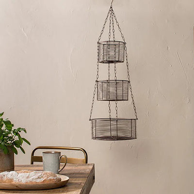 3 tier wire hanging basket against taupe wall next to wooden table with bread loaf, mug and green plant
