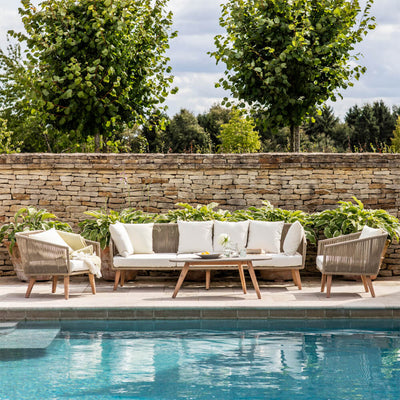 Idylic image of a Colwell three piece seating set with cream cushions beside a blue swimming pool with a stone wall and trees behind