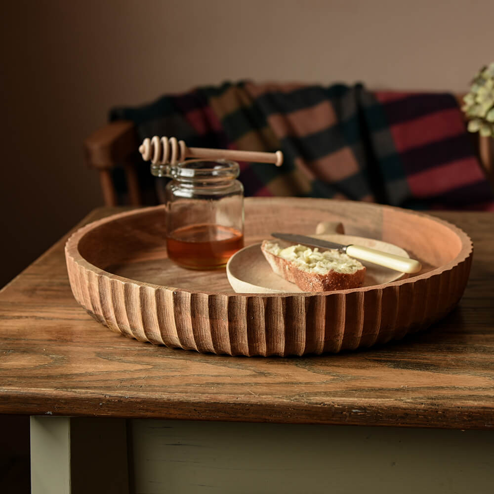 Carved edge to a large round flat wooden bowl with bread and honey inside on a rustic kitchen table
