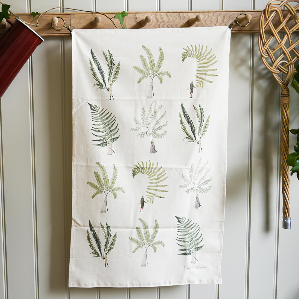 Tea towel with fern pattern hanging on a wooden rack against a white paneled wall.