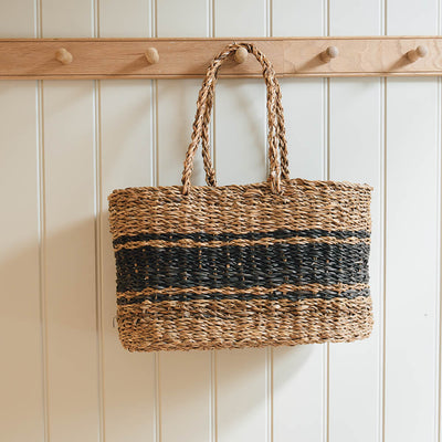 Woven bag with black stripes hanging on a wooden coat rack against a white paneled wall.