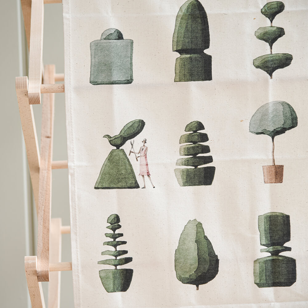 Decorative tea towel with green topiary trees on a white background, hanging on a wooden airer