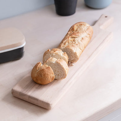 Alderwood long serving board with a white baguette sliced up sitting on a wooden worktop.