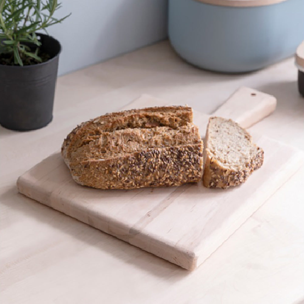 Alderwood square serving board with a loaf of wholegrain bread on a wooden worktop against a pale blue wall. 