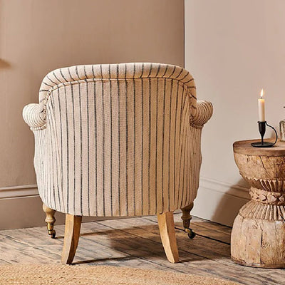 Back view of a black and white striped linen armchair with mango wood legs placed in the corner of a room next to a rustic wooden table with a lit candle.