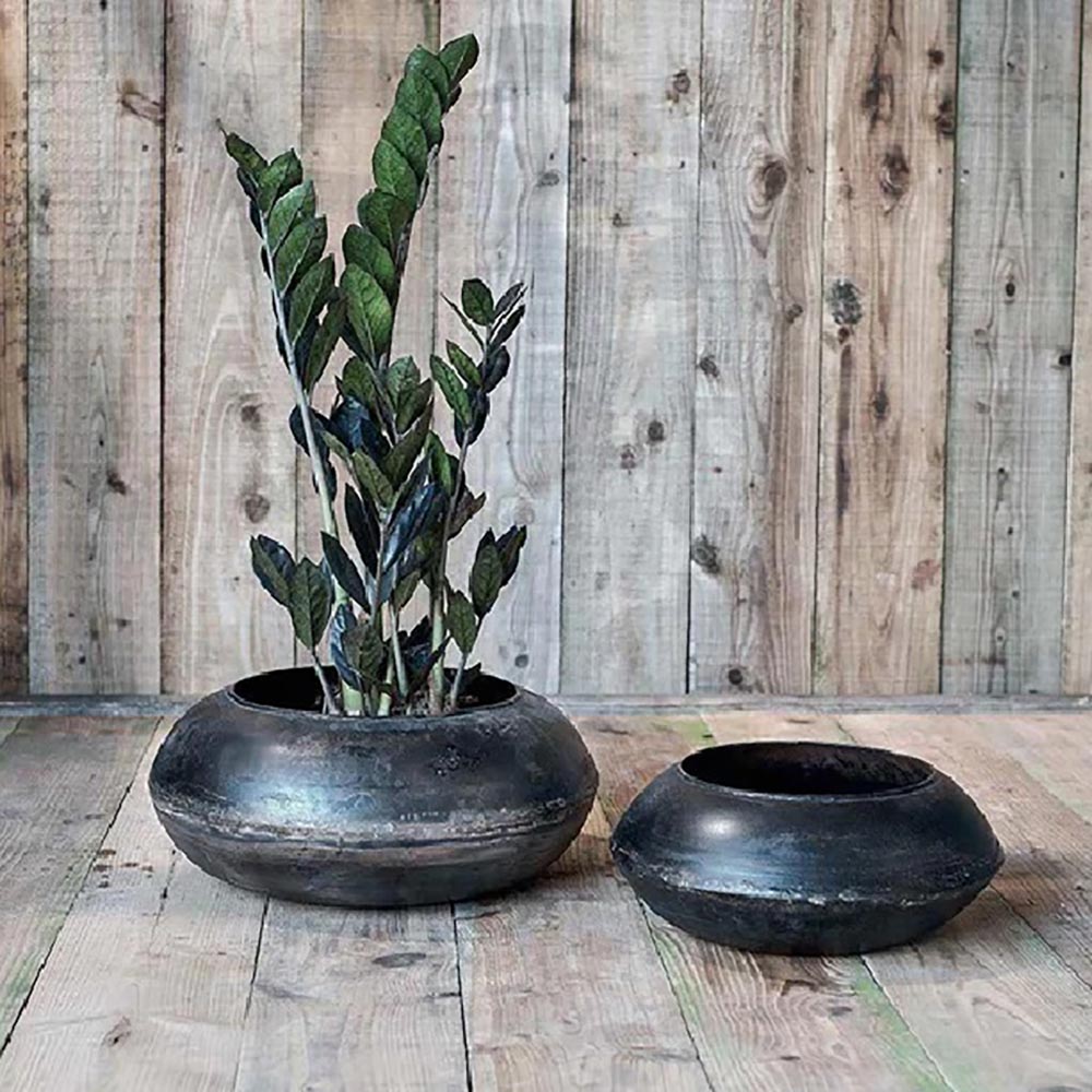 Black reclaimed iron planters in a flat round design on a wooden floor with wood panelled wall. One contains a plant while the other is empty.