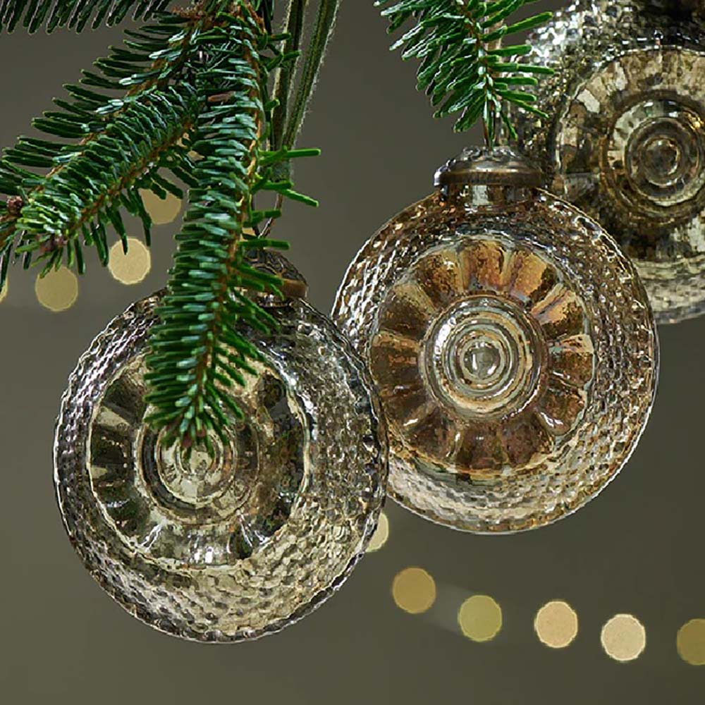 Close up of vintage style glass baubles in an antique gold and grey finish with a shell-like design hanging from a christmas tree branch against a dark green background