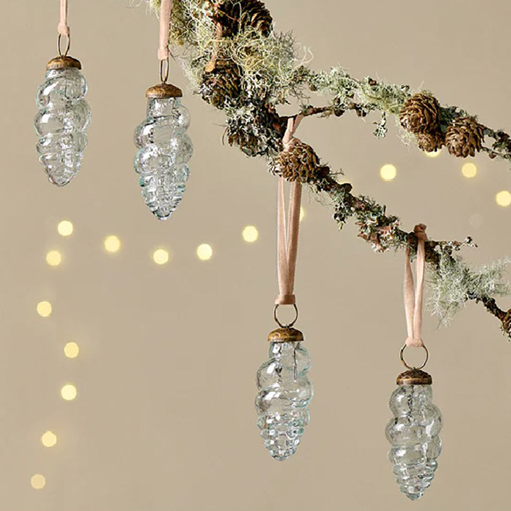 Close up of a set of 4 clear glass teardrop baubles in a shell-like design with light pink velvet ribbon hanging from a branch against a pale yellow background