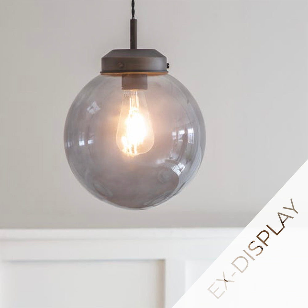 Round globe glass pendant light in a smoky grey colour with antique brass fittings illuminted against a white wall with a watermark and ex display text in the corner.
