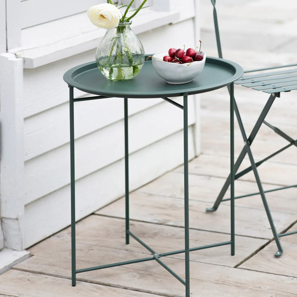 Small round metal folding table in forest green with a green glass vase and bowl of cherries on top