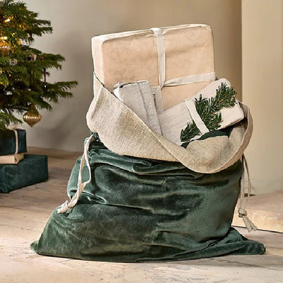 Forest green velvet christmas sack with a jute linen band at the top. Placed on a wooden floor next to a christmas tree with presents hanging out of the top.