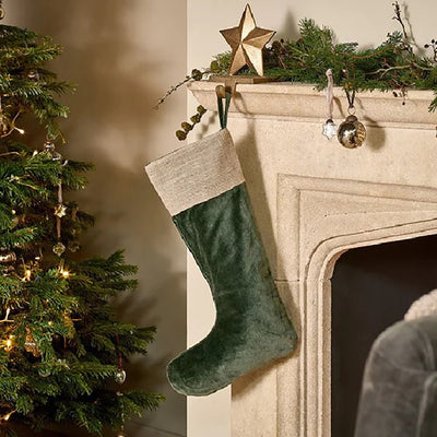 Forest green velvet christmas stocking with jute band around the top. Hanging from stone fireplace on a gold star hook next to a christmas tree with gold baubles.