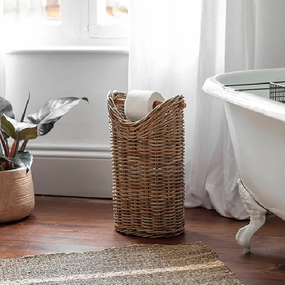 Rattan toilet roll holder with storage basket underneath placed on wooden floor next to white enamel bath with window and white curtains behind 