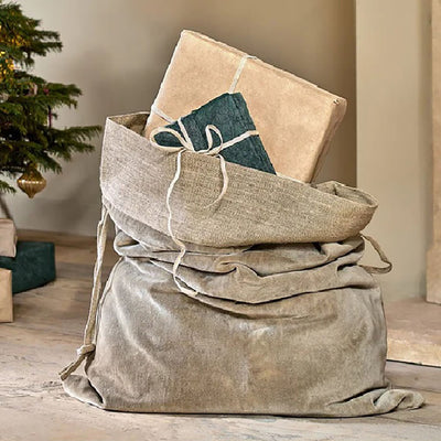 Light grey velvet christmas sack with a jute linen band at the top. Placed on a wooden floor next to a christmas tree with presents hanging out of the top.