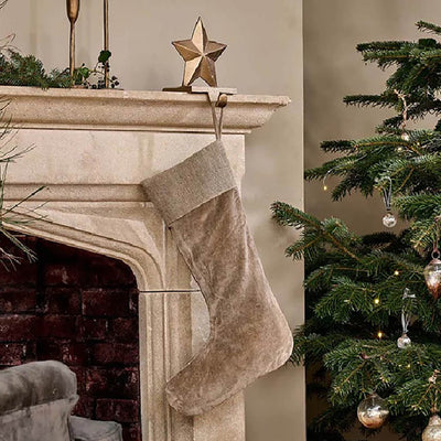 Light grey velvet christmas stocking with jute band around the top. Hanging from stone fireplace on a gold star hook next to a christmas tree with gold baubles.