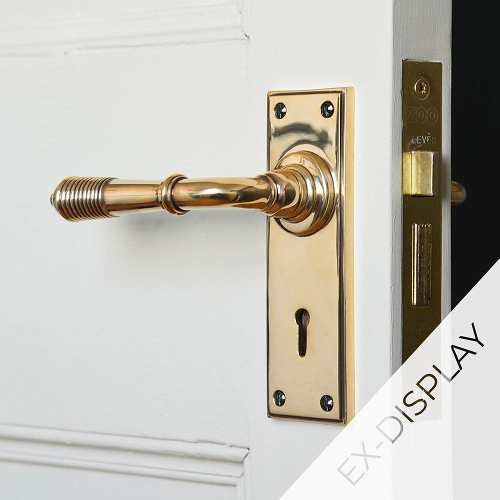 Polished antique brass reeded lever handles with a rectangular backplate and keyhole on a white door with a watermark and ex display text in the corner