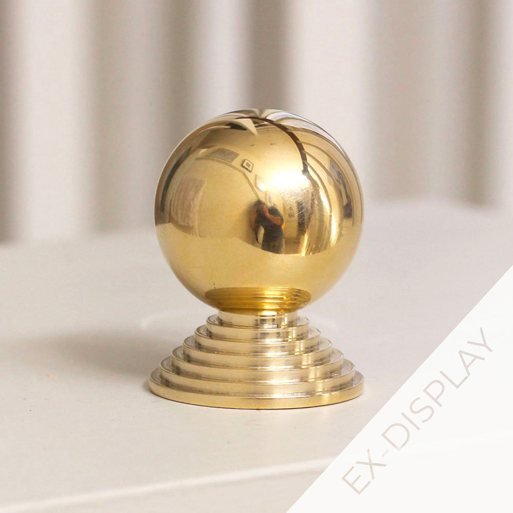 Polished brass round ball cabinet knob with a stepped backplate on a pale pink surface with a watermark and ex display text in the corner