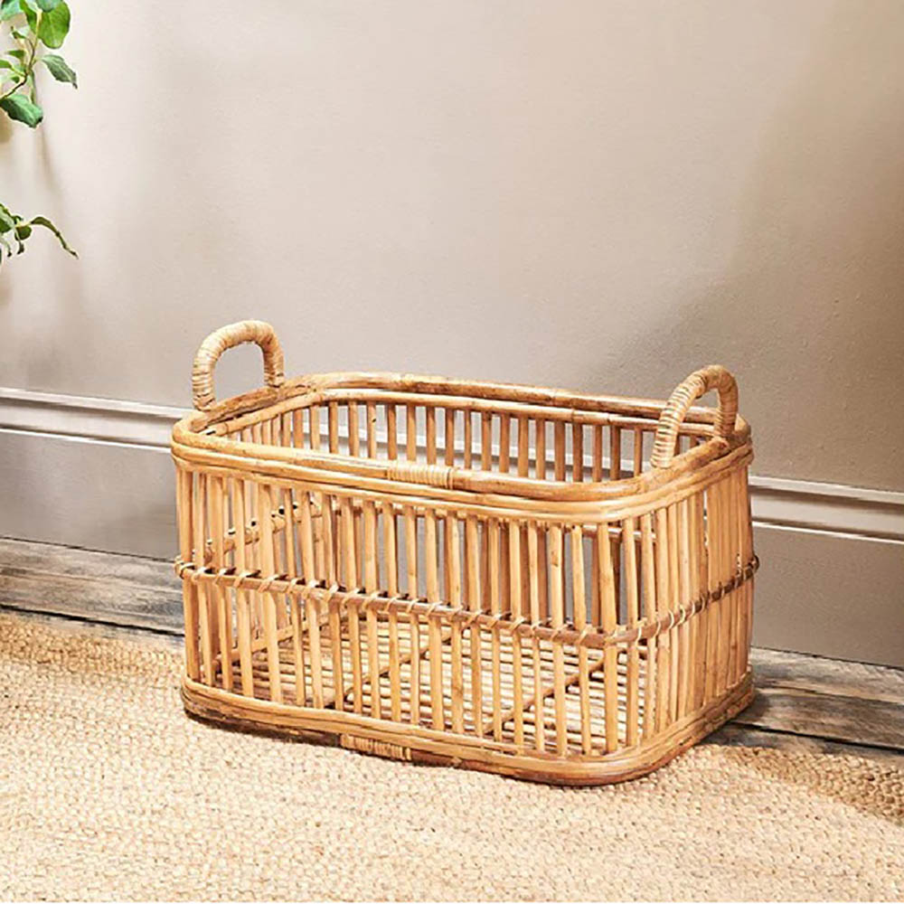 Rectangular rattan laundry basket with curved handles against a taupe wall on wooden floor