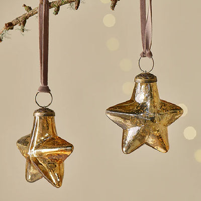 Close up of rustic gold glass star baubles with pink velvet ribbon hanging from a tree branch against a pale yellow background