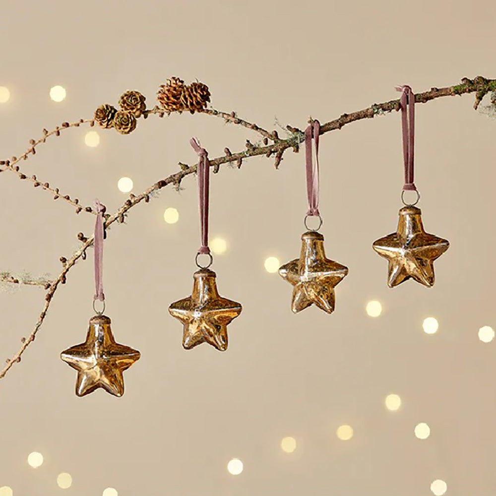 Set of 4 rustic gold glass star baubles with pink velvet ribbon hanging from a tree branch against a pale yellow background