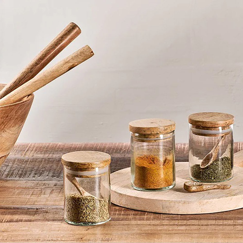 Set of 3 glass spice jars with mango wood lids and wooden spoons filled with herbs and spices on wooden table against pale background with wooden serving bowl to left of picture