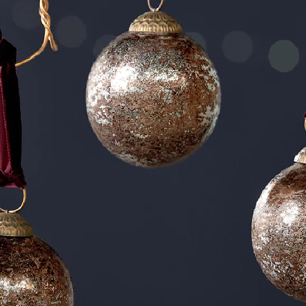 Close up of a round glass bauble in a rustic antique burgundy finish with a deep red velvet tie hanging from copper wire against a dark blue background