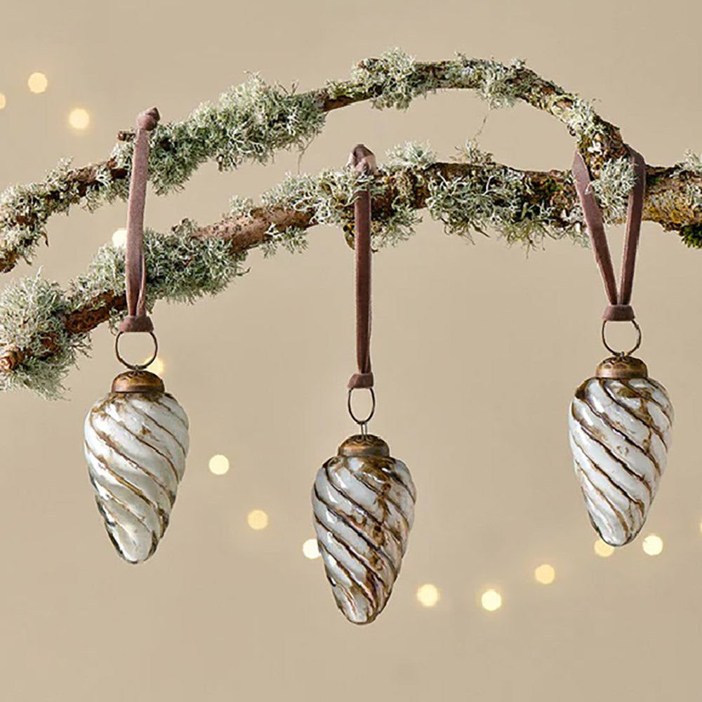 Set of white and gold teardrop glass baubles in a shell like design with pink velvet ribbon hanging from a tree branch against a pale yellow background