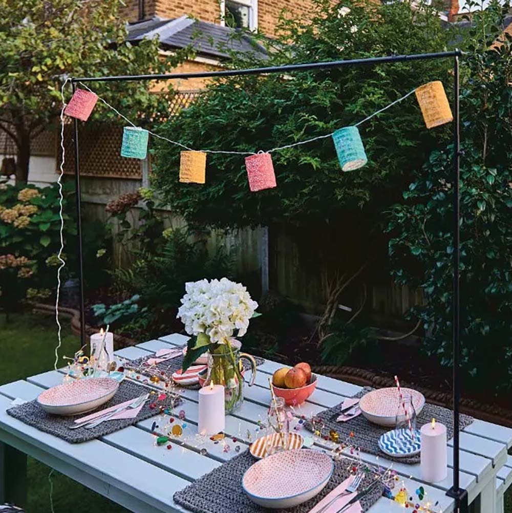 Set of 6 paper lanterns in teal, yellow and pink strung over an outdoor table, hanging from a black table frame