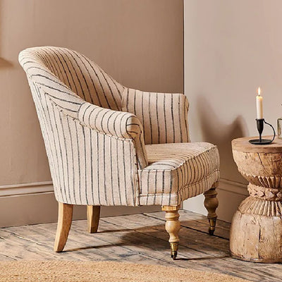 Side view of a black and white striped linen armchair with mango wood legs placed in the corner of a room next to a rustic wooden table with a lit candle.