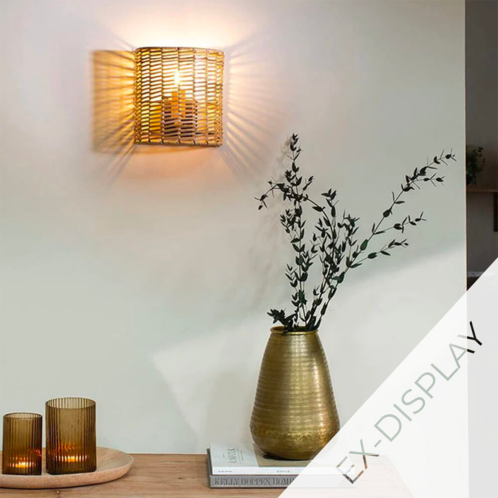 Small noko rattan wall light illuminated on an off-white wall with a brass vase an greenery below. A watermark and ex display text are in the corner.