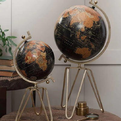 Small and large decorative world globes with solid brass tripod stands sat on a round wooden table against a pale background. The world maps feature black and burnt orange map images
