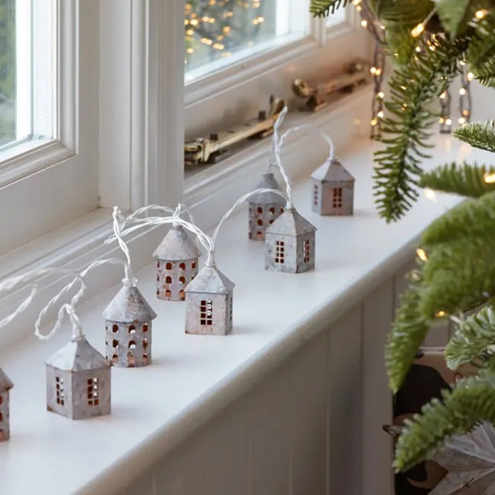 Warm white LED string lights featuring rustic white metal village type house on a white wire. Placed on a white windowsill next to a christmas tree
