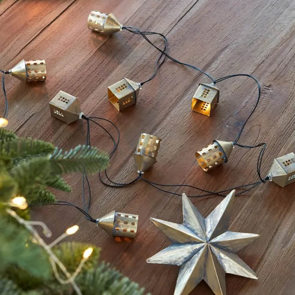 Warm white LED fairy lights featuring gold metal village houses on a black wire displayed on  a wooden table next to  a christmas tree