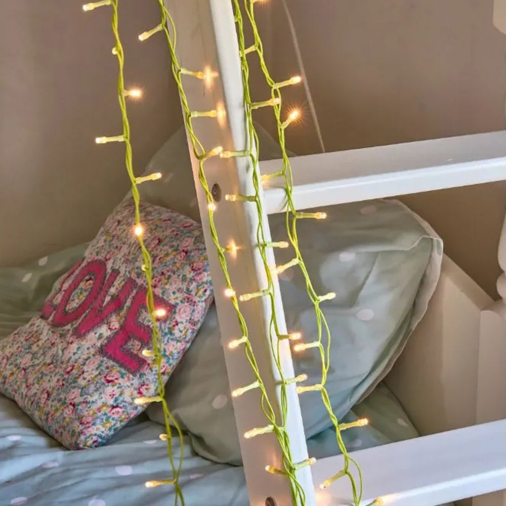 Warm white fairy lights with a green cable hanging from a bunk bed ladder with a bed and cushions in the background