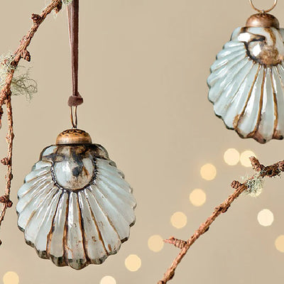 Close up of white and gold glass shell baubles with pink velvet ribbon hanging from a tree branch against a pale yellow background