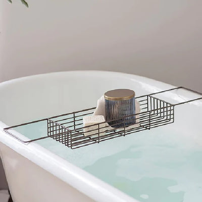 Iron wire bath caddy with an antique brass finish resting on an enamel bath filled with water with a glass jar and bar of soap in the tray