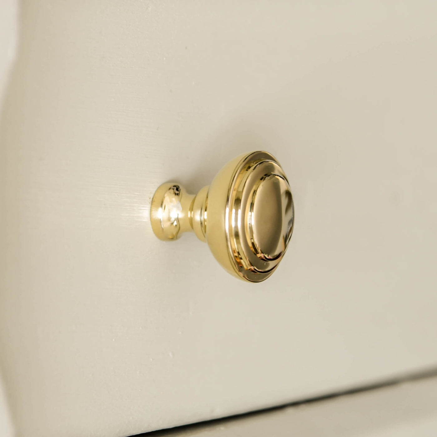 Round brass cabinet knob with ribbed detailing on the front seen on an off white painted drawer