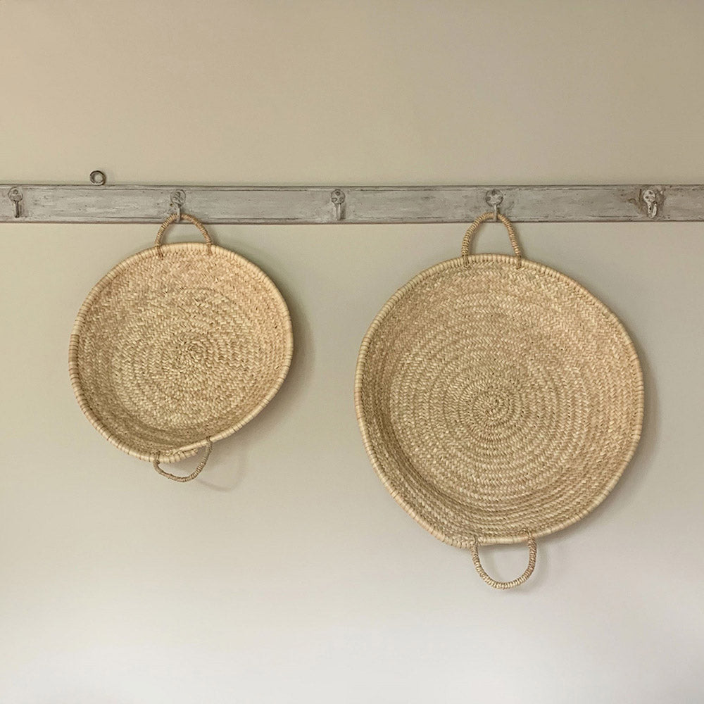 Round palm leaf trays in a small and large size hanging next to each other on a hook rail against an off-white wall