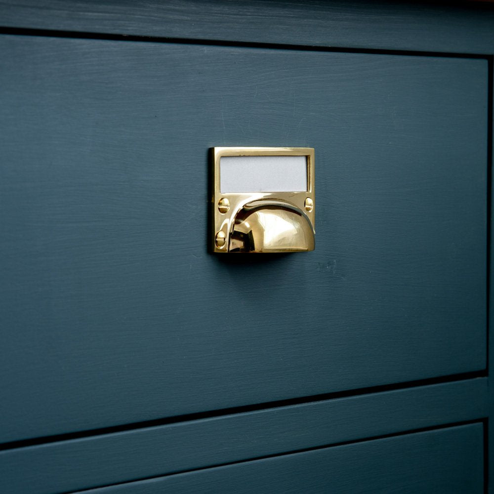 A Hooded Drawer Pull with Traditional Card Frame in Polished Brass on a dark blue drawer